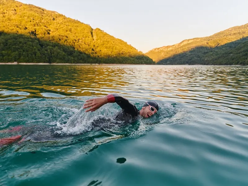 A triathlete in a wetsuit and goggles swimming freestyle across a mountain lake at sunset