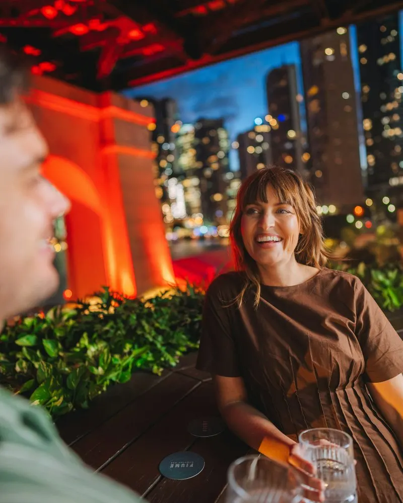 A smiling woman with a drink at a busy rooftop bar at night, city towers lit up behind her, chatting with a man in the foreground