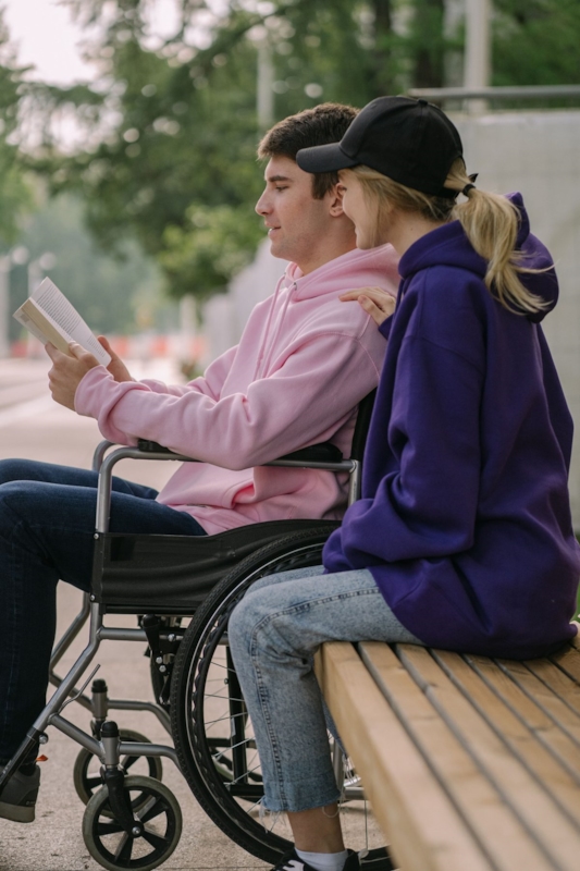 A man in a wheelchair reading aloud while a woman sits close beside him on a bench
