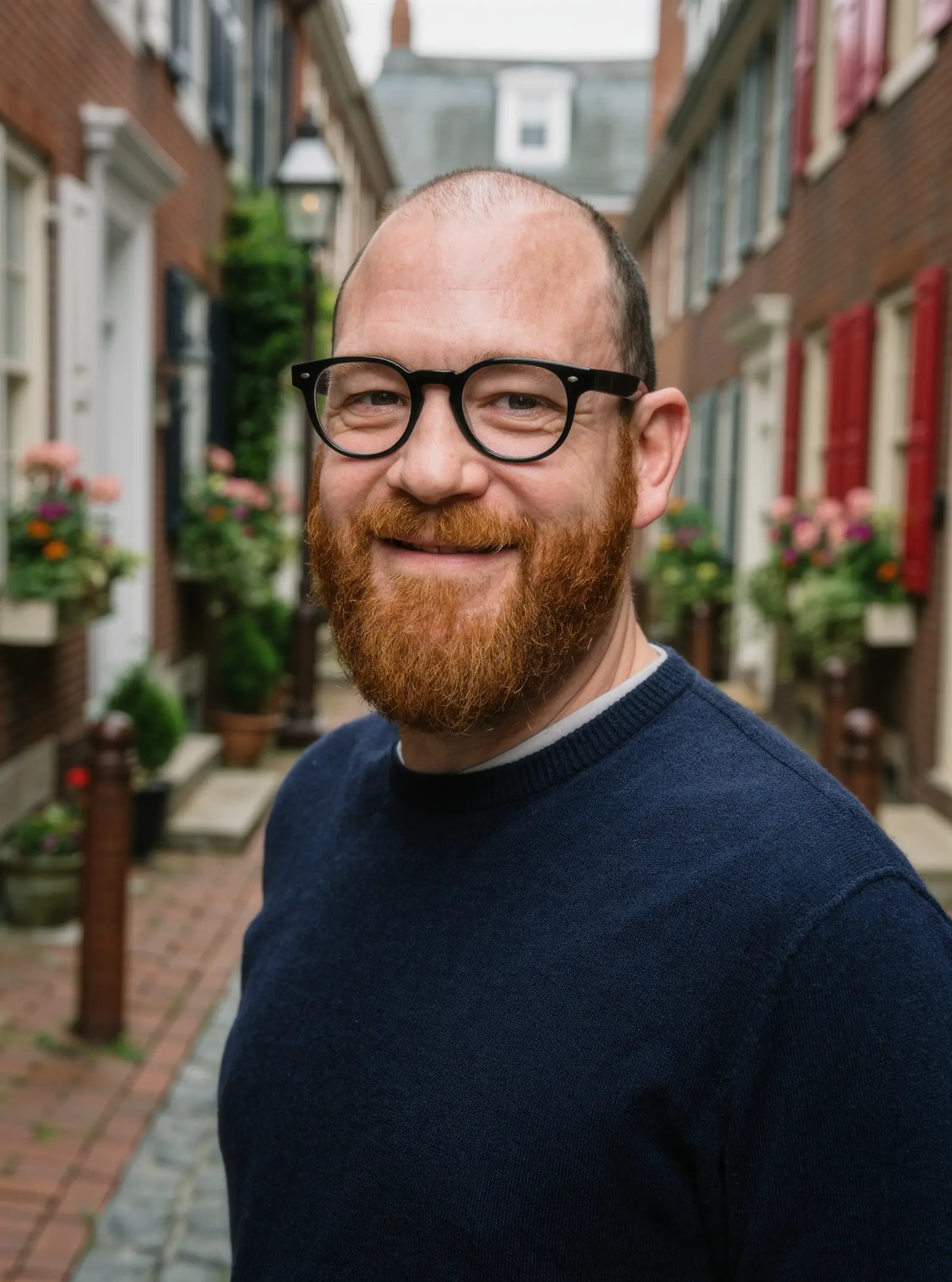 A smiling man with glasses and a red beard on a narrow brick lane of rowhouses with flower boxes