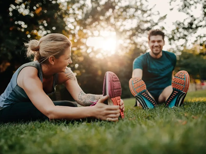 A woman and man laughing as they stretch together on the grass in a sunny park