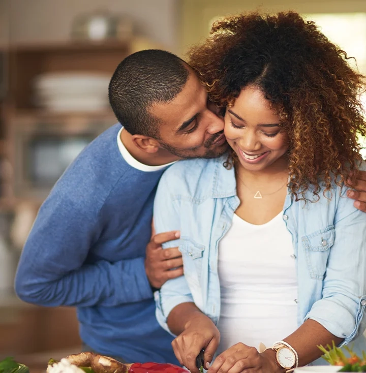 A couple cooking together, him kissing her cheek as she laughs