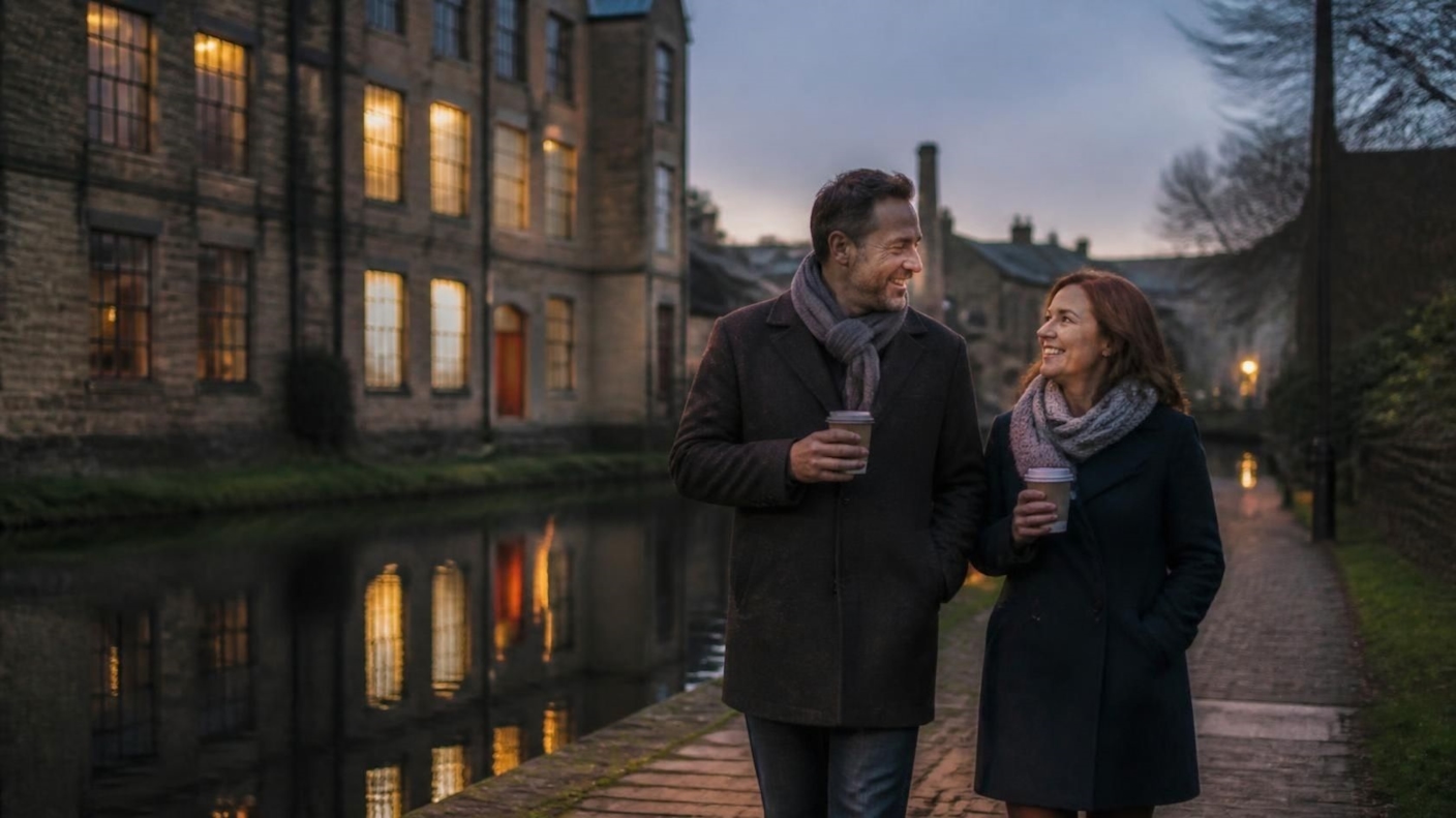 A couple in winter coats walking along a canal towpath at dusk past old stone mills