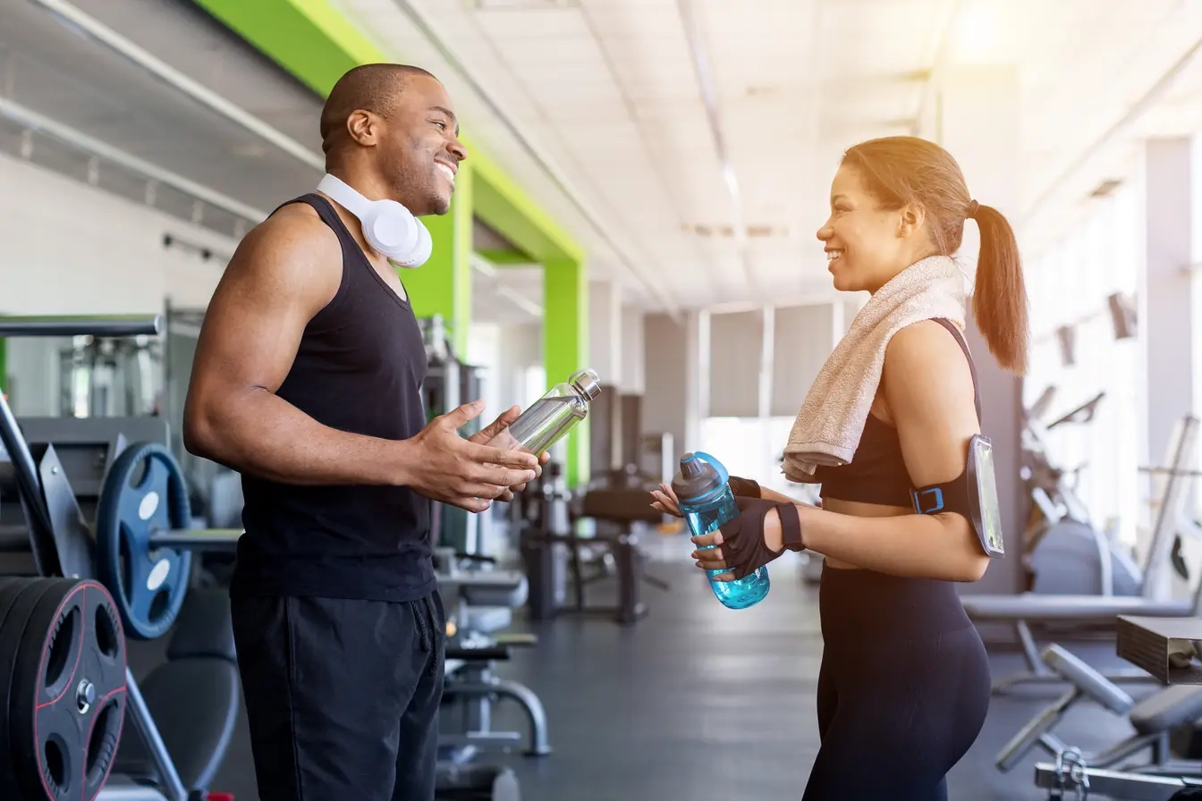A smiling man and woman chatting with water bottles between sets in a bright gym