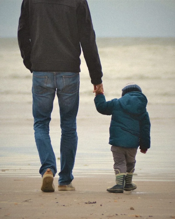 Father and small child walking hand in hand on a beach, seen from behind
