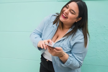 A woman in a denim shirt laughing at something on her phone against a mint green wall