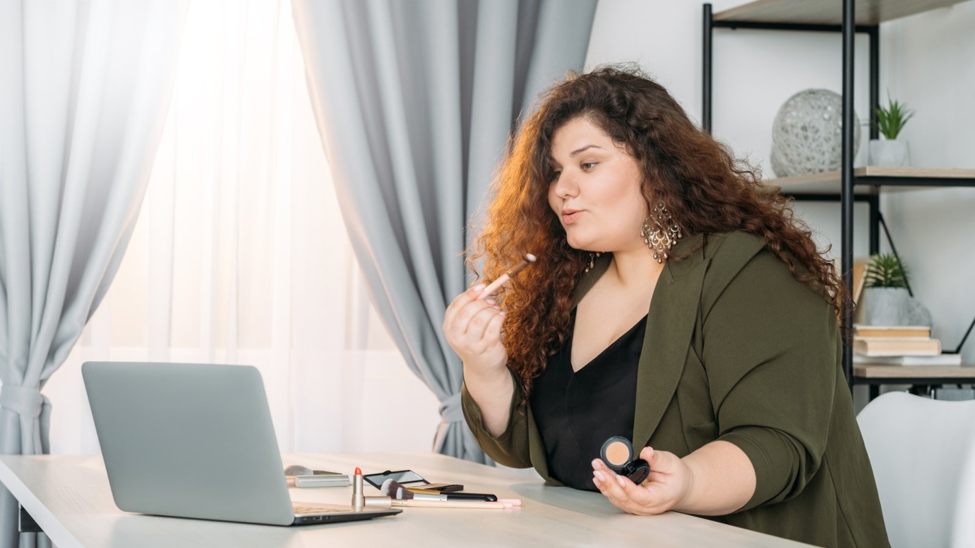 A woman applying makeup at her desk, getting ready with her laptop open