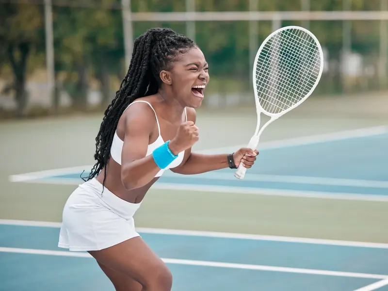 A woman with a tennis racket cheering after winning a point on an outdoor court