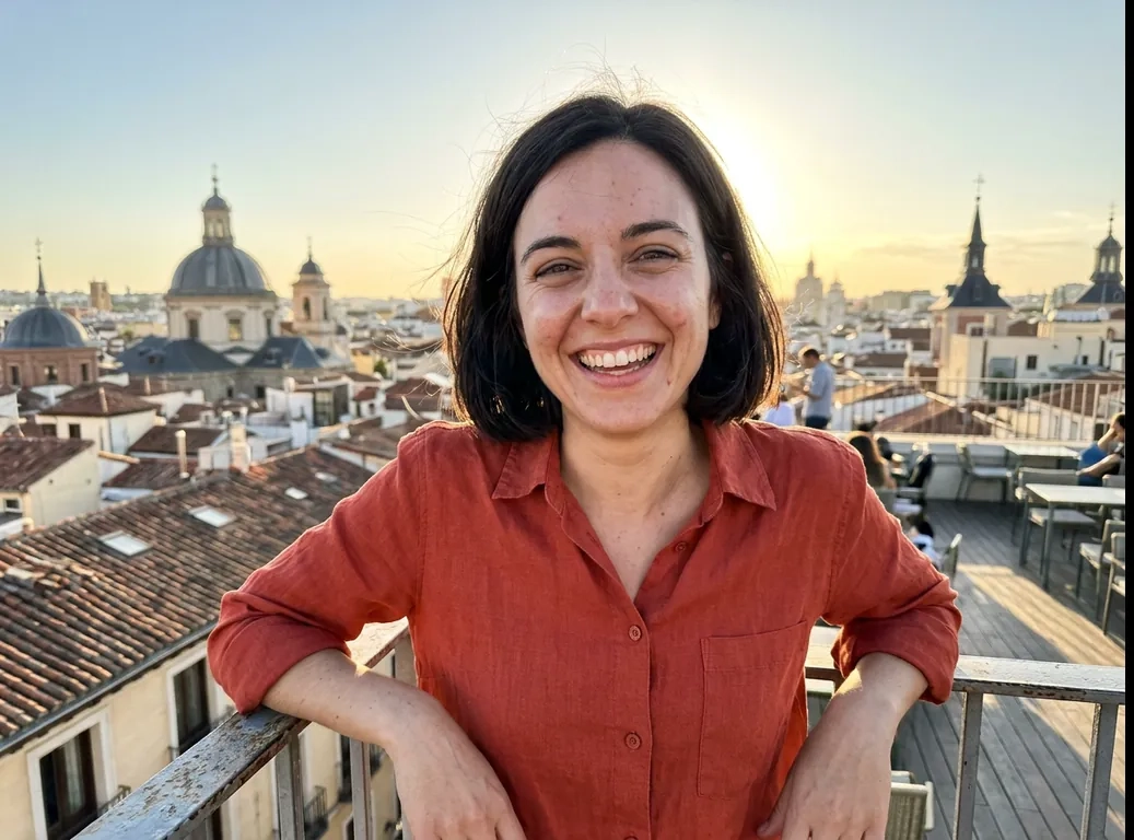 Una mujer sonriente con media melena oscura y una camisa naranja se apoya en la barandilla de una azotea de Madrid al atardecer, con cúpulas y tejados detrás (imagen generada por IA)