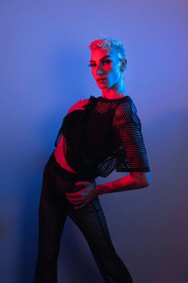 una persona de pelo corto platino bajo una luz azul y rosa, con camiseta de rejilla