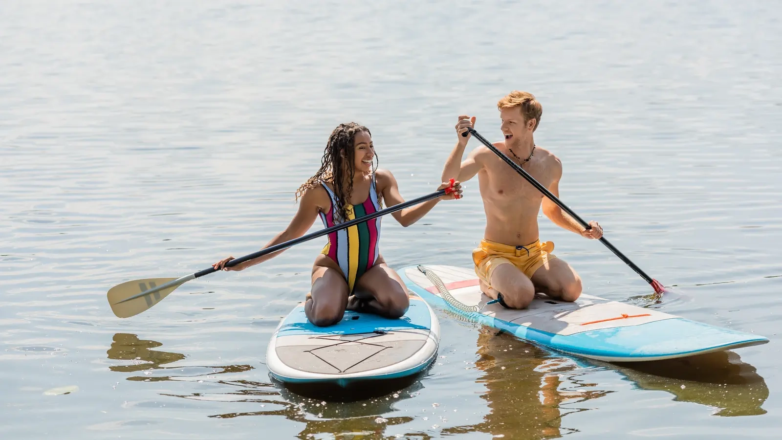 A laughing couple kneeling side by side on paddleboards on a calm, sunlit lake
