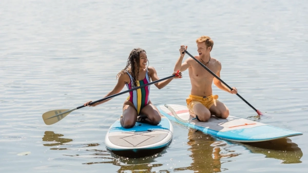 A laughing couple kneeling side by side on paddleboards on a calm, sunlit lake