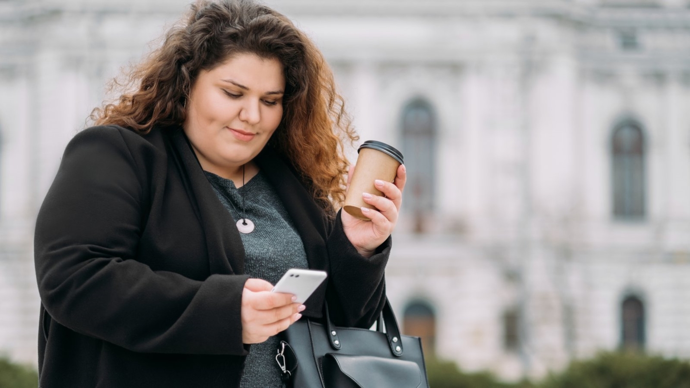 A woman checking her phone while holding a coffee, walking through the city