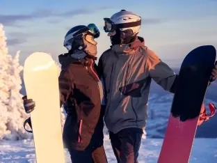 A couple of snowboarders standing close together holding their boards on a snowy slope at sunset