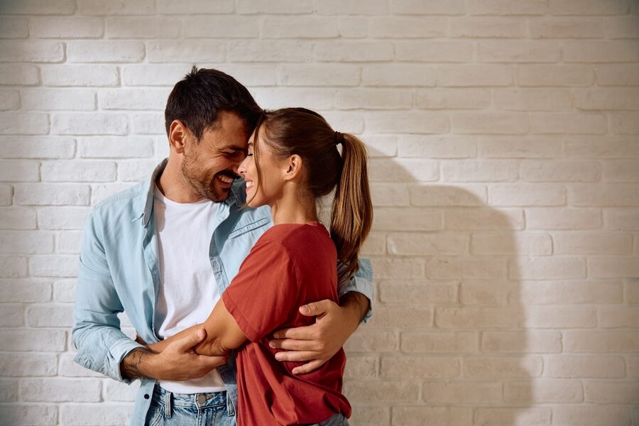 Couple embracing and smiling at each other against a white brick wall