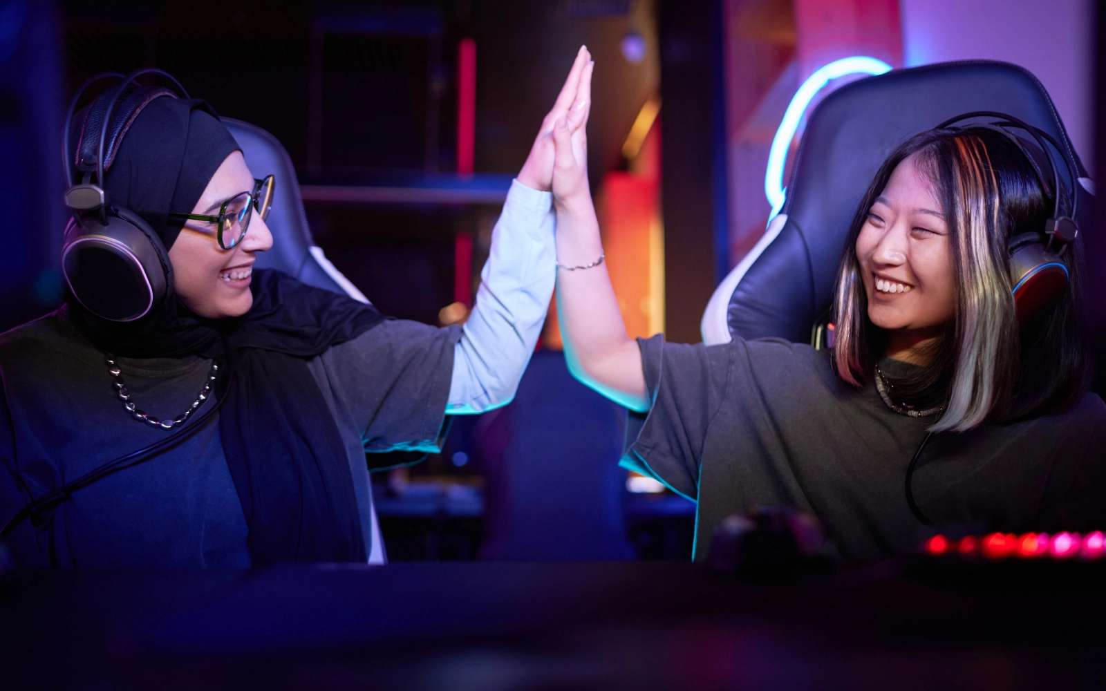 Two friends in headsets high-fiving at their gaming chairs