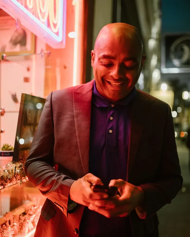 A smiling bald man in a blazer checking his phone outside a neon-lit window at night