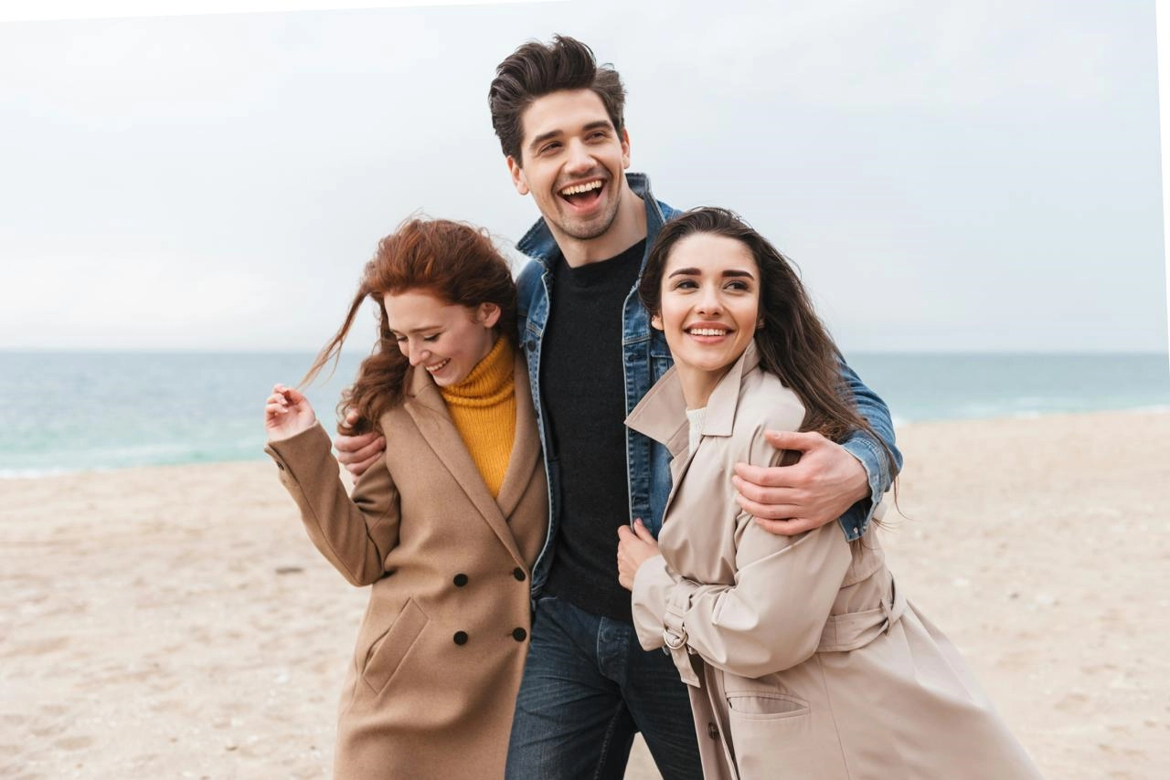 Trío sonriendo abrazado en la playa, con abrigos de entretiempo