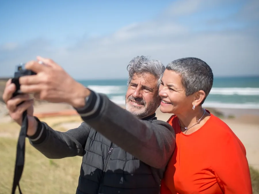 A smiling couple with grey hair taking a selfie with a camera on a sunny beach