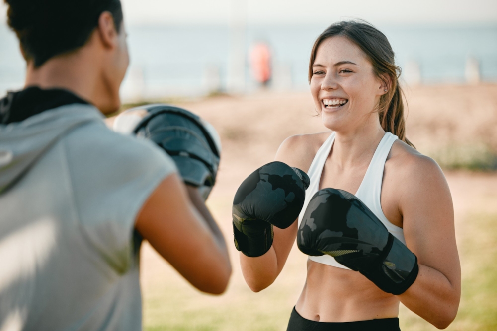 A laughing woman in boxing gloves training with a partner holding pads outdoors