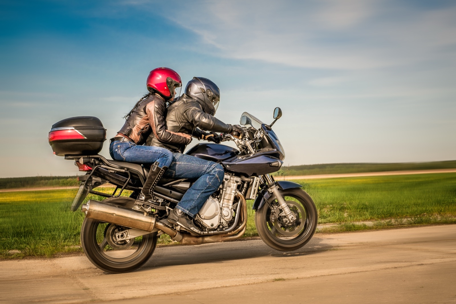 A rider and pillion in helmets riding two-up along an open country road on a sunny day