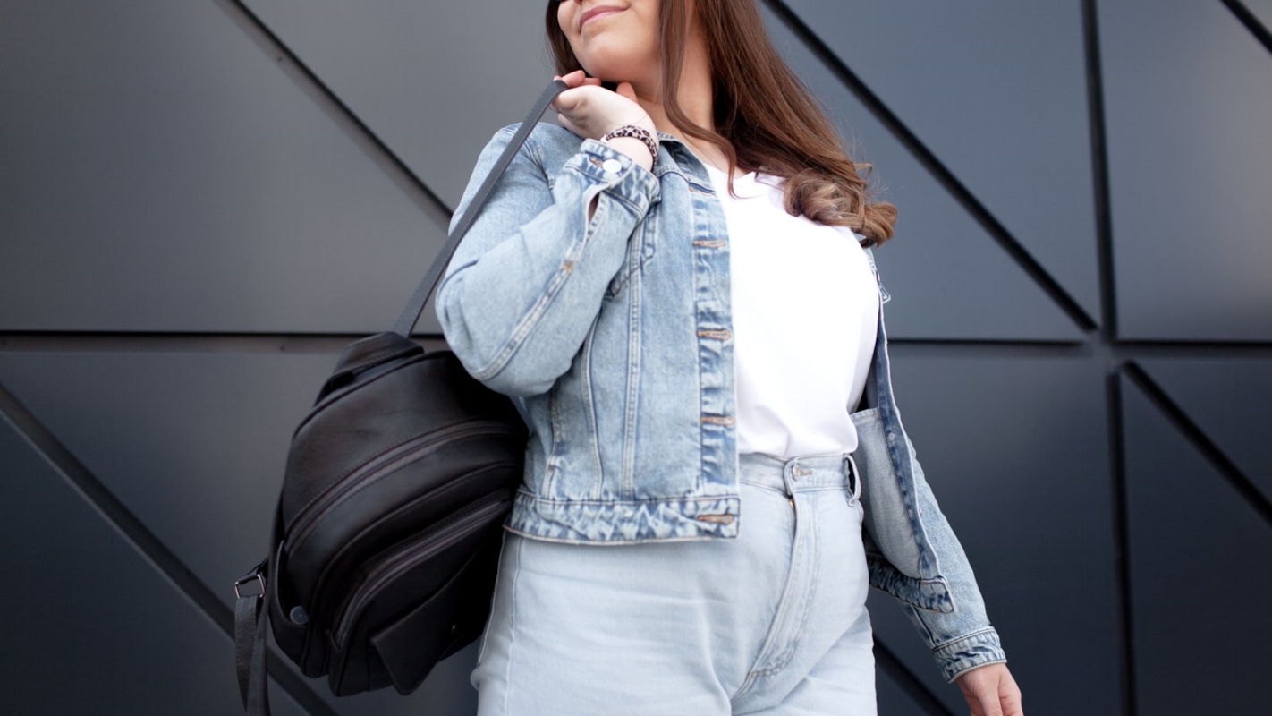 A woman walking outdoors in a denim jacket, carrying a backpack