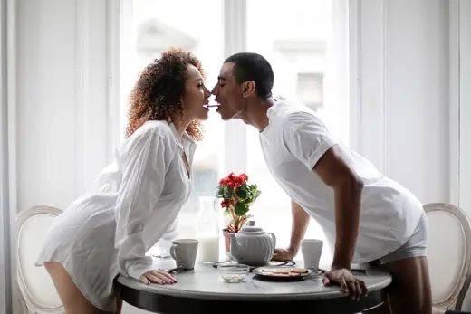 A couple leaning across a breakfast table to kiss in bright morning light