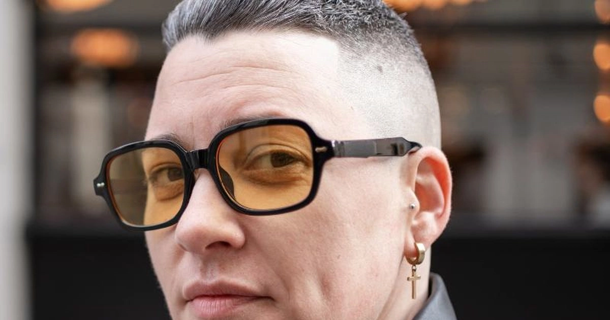 A woman with short hair and amber-tinted glasses looks towards the camera