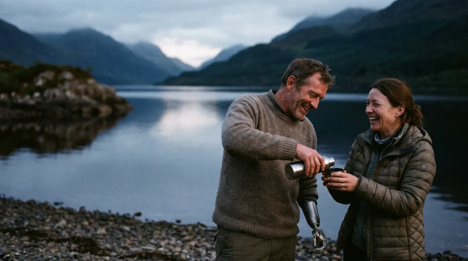 A couple laughing by a Highland loch at dusk as the man, who has a prosthetic arm, pours tea from a flask
