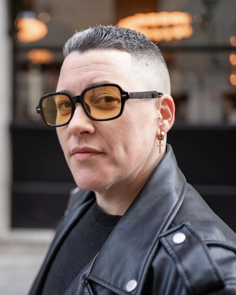A woman in a leather jacket stands on a city street, looking towards the camera