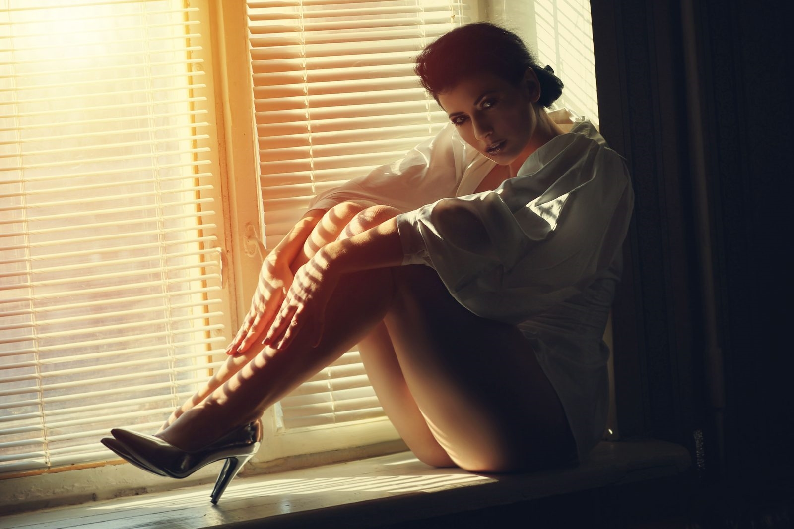 Sexy mature woman in a white shirt and heels sitting on a sunlit windowsill