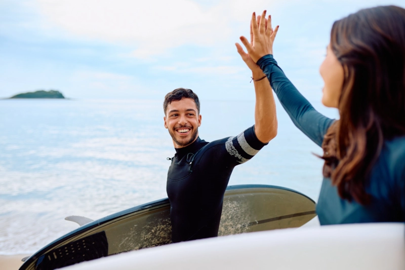 A smiling man and woman in wetsuits high-fiving on the beach with their surfboards
