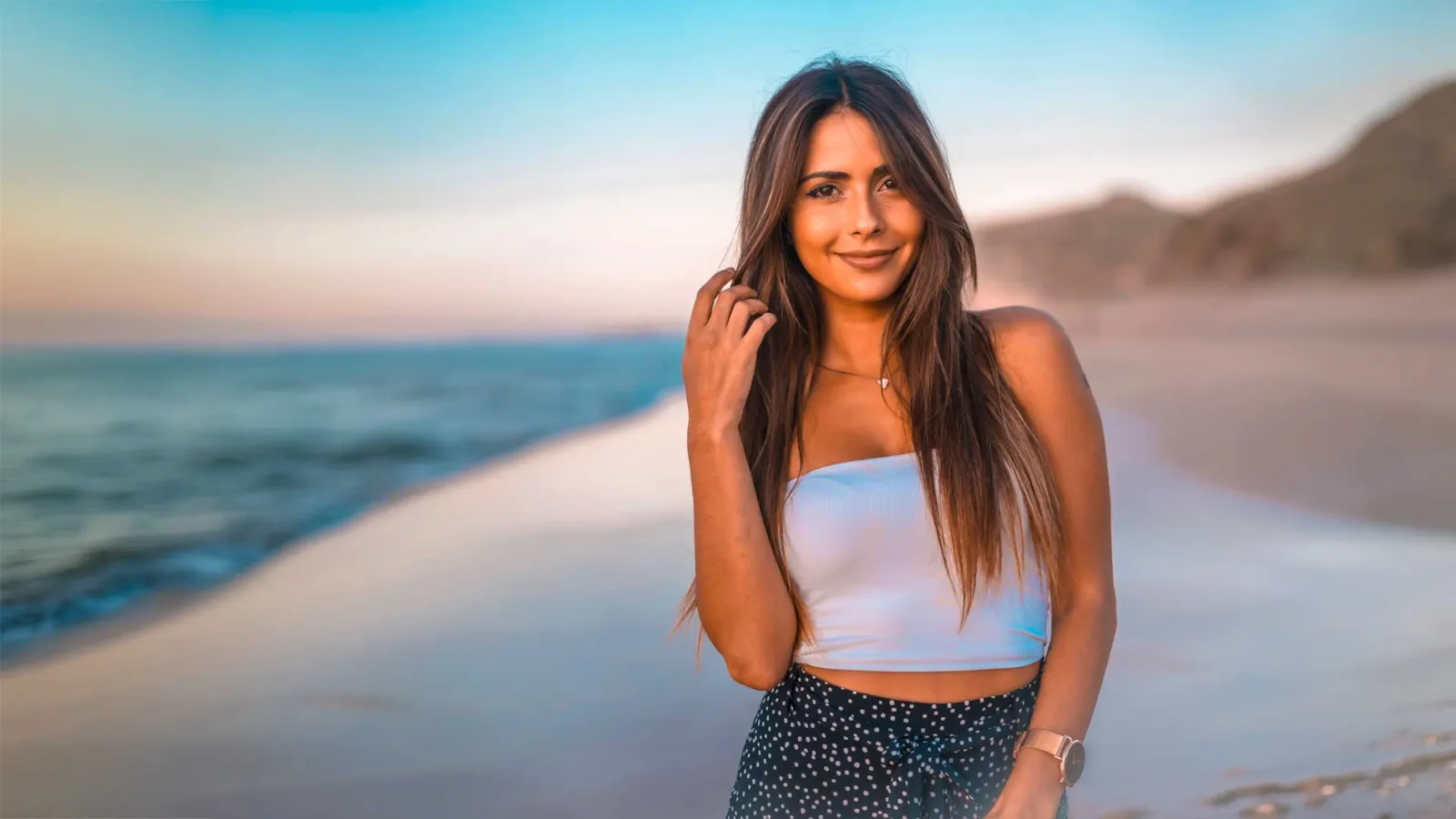 A smiling woman with long brown hair standing on a beach at sunset