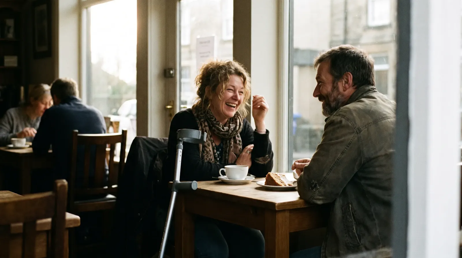 A couple laughing over tea and cake at a café window table; the woman has cerebral palsy and her forearm crutches rest against the table