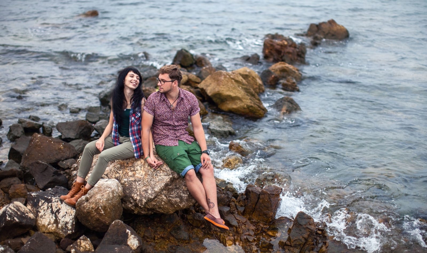Young couple sitting on rocks by the sea, holding hands and smiling