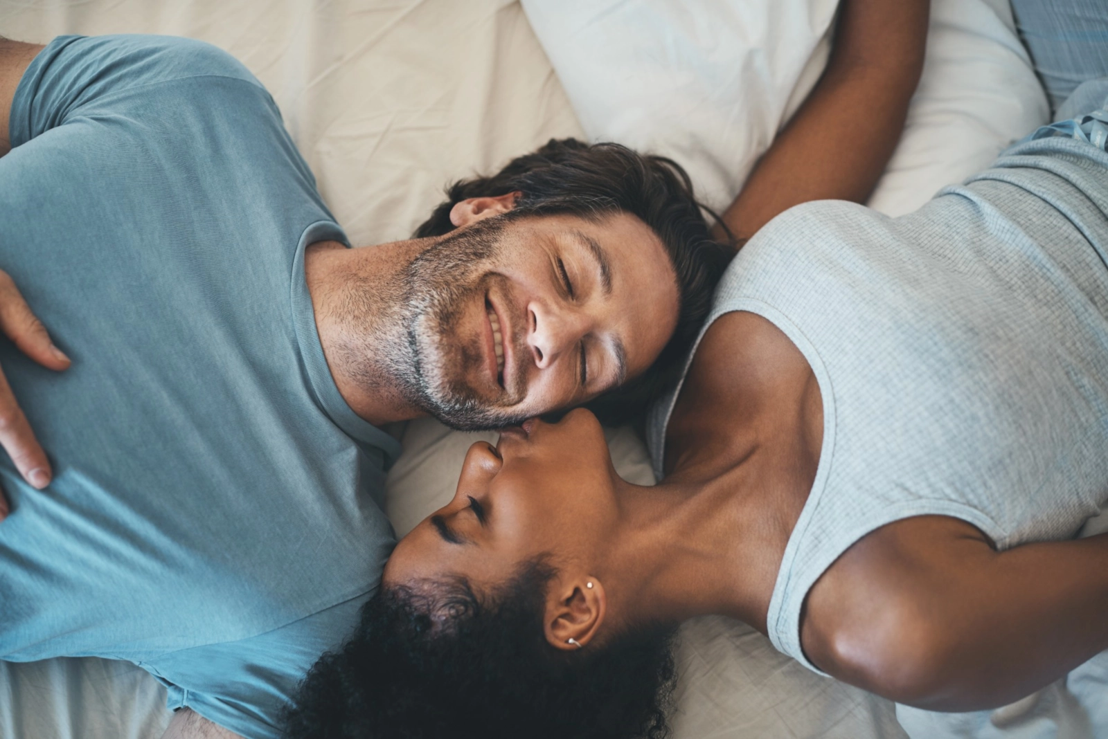 A smiling couple lying head to head on a bed, she kisses his cheek