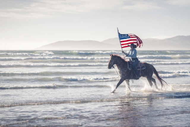 A rider on horseback splashing through the surf with the American flag flying