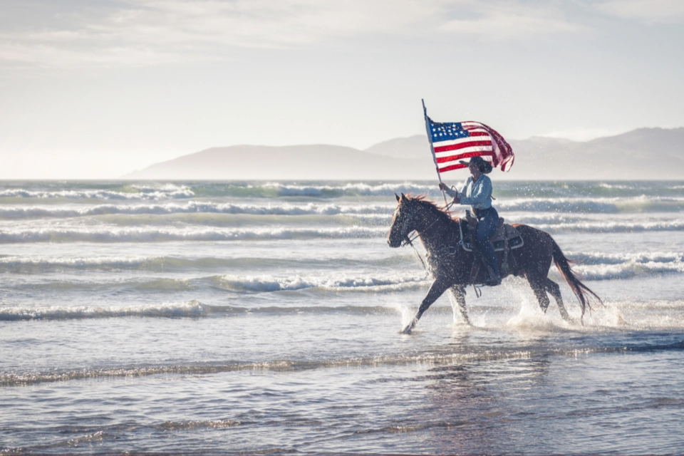 A rider on horseback splashing through the surf with the American flag flying