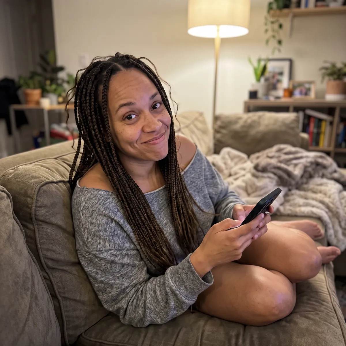 A woman with long box braids in an off-the-shoulder grey sweatshirt curls up on a sofa with her phone and gives the camera a playful grin (AI-generated image)