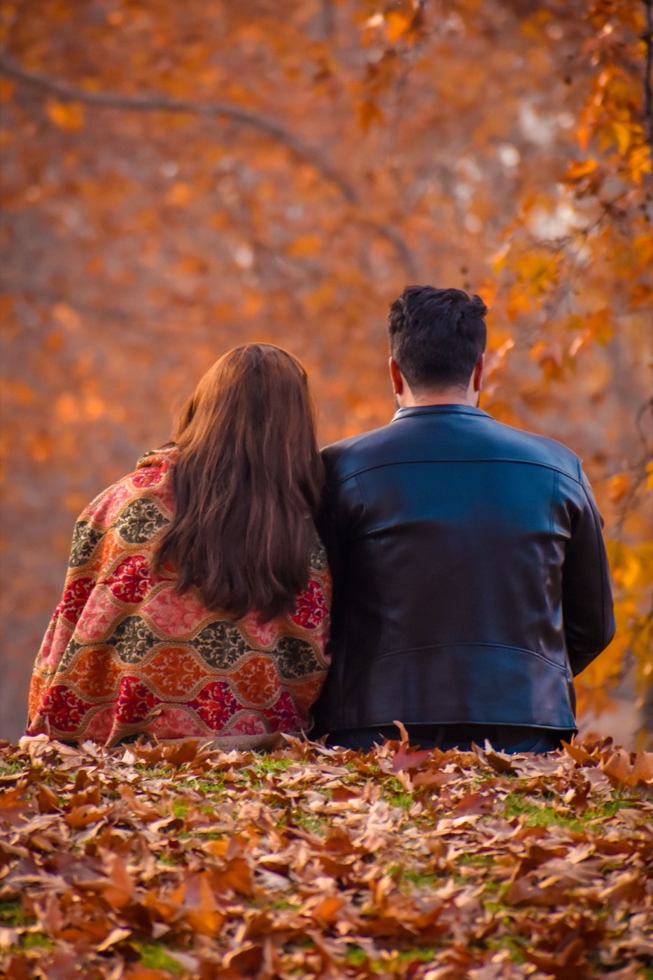 A man in a dark jacket and a woman in a patterned shawl sit together on autumn leaves facing a forest of orange trees