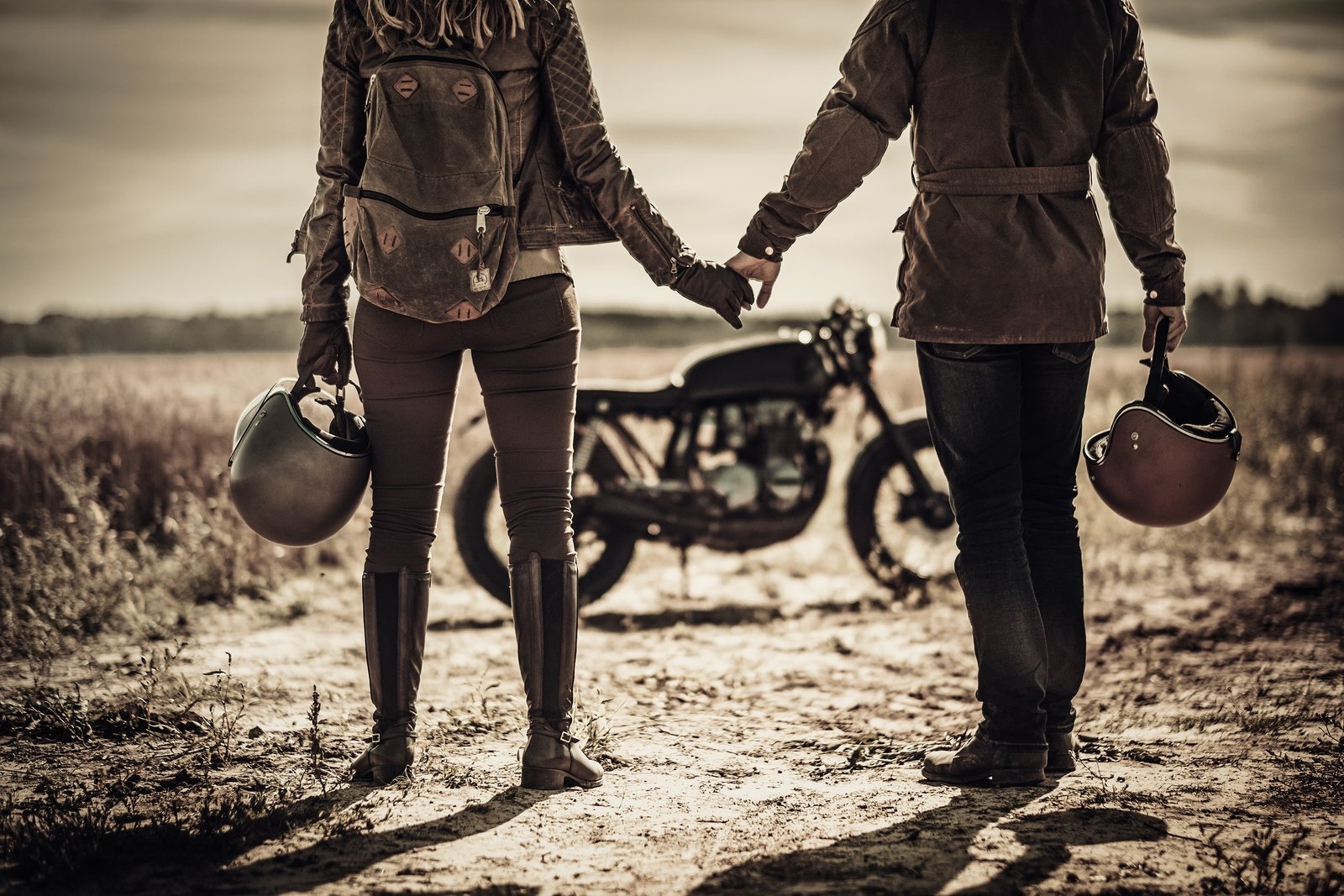 A couple holding hands and carrying their helmets beside a cafe racer motorcycle in a sepia-toned field