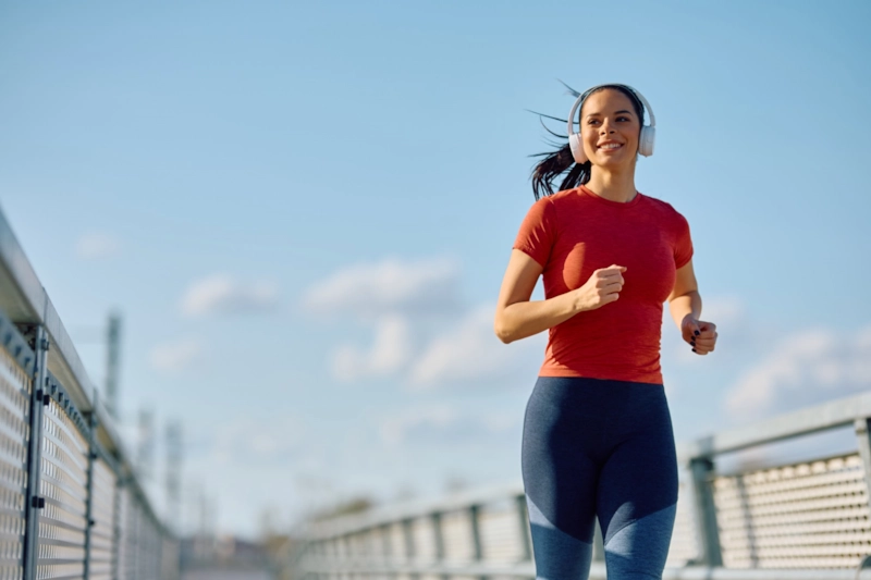 A smiling woman in headphones jogging across a bridge under a blue sky