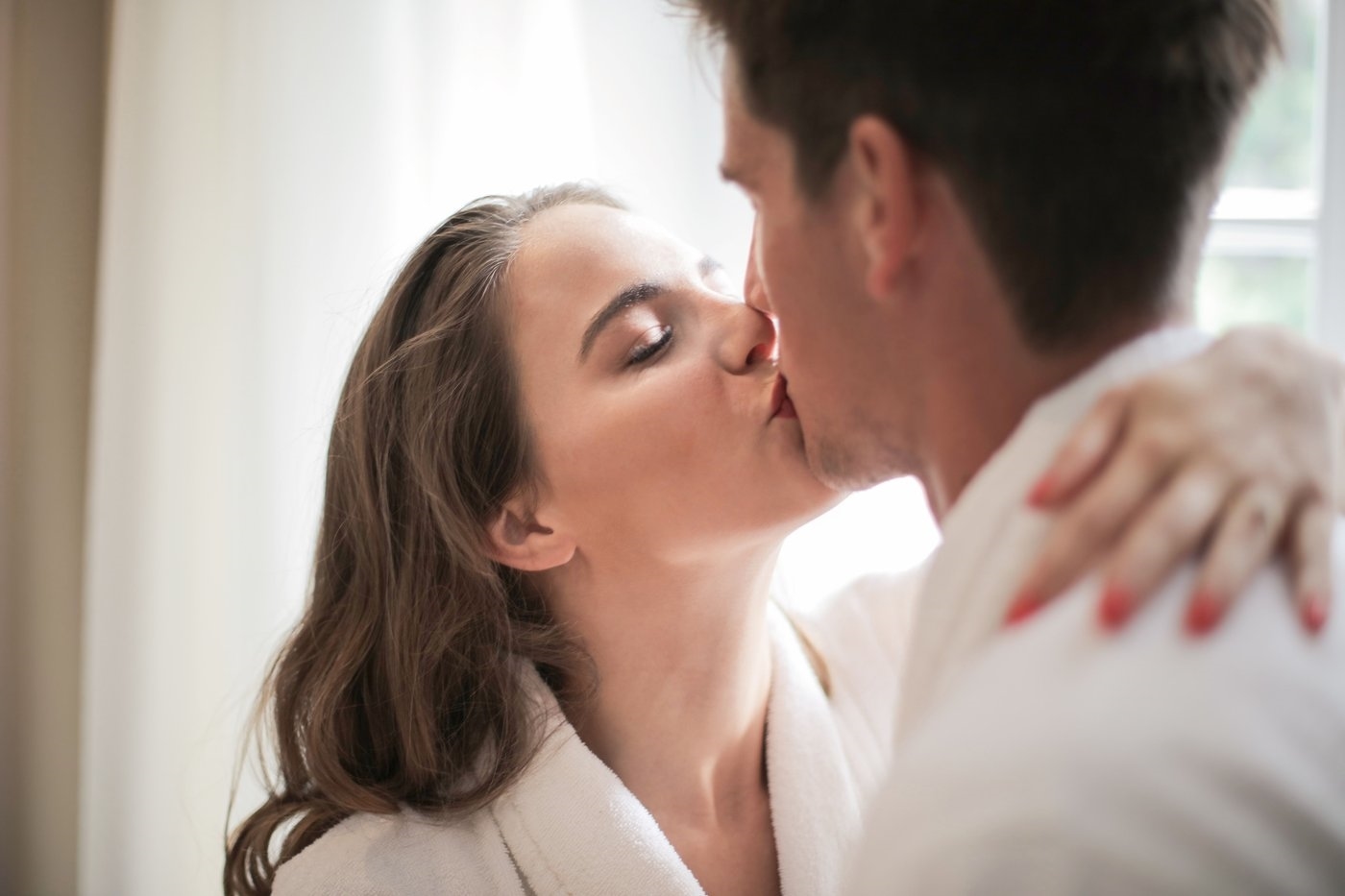 A woman with long dark hair kissing a man in a white shirt in a bright room, illustrative photo of models