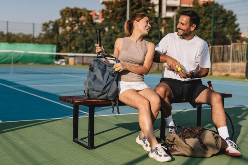 A woman and man laughing together on a bench beside an outdoor tennis court