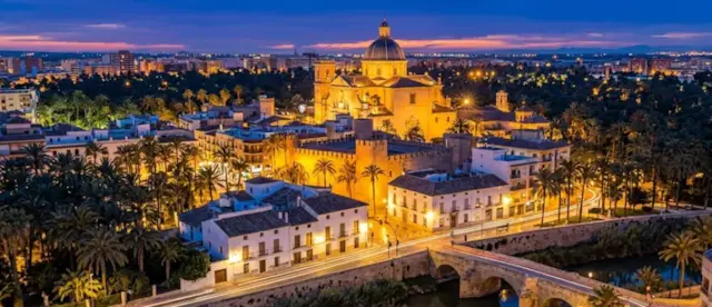 Basílica de Santa María de Elche iluminada al anochecer entre palmeras