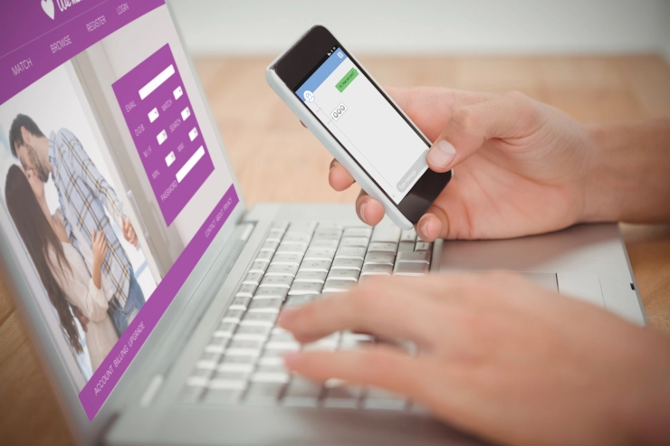 A man holding a mobile phone while typing on a laptop at a desk, with a dating app on screen