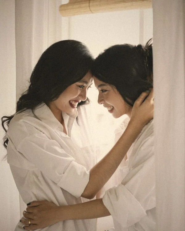 Two women in white shirts smiling at each other by a window