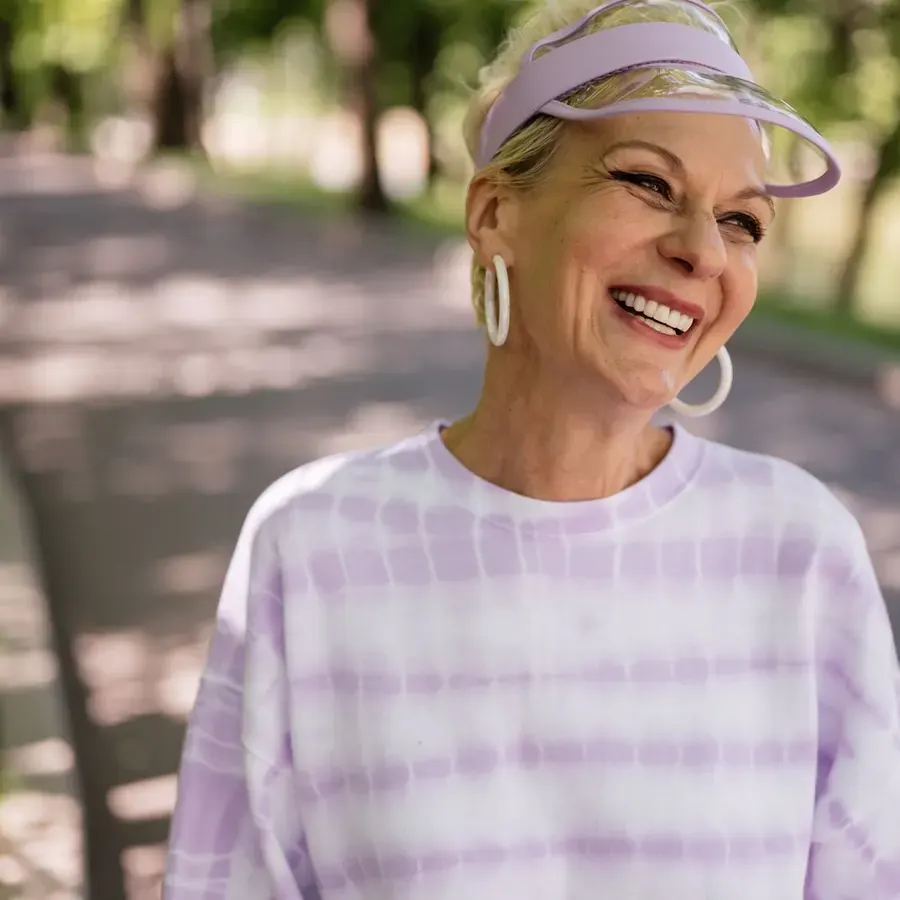 A woman in a sports visor and hoop earrings laughing on a tree-lined path