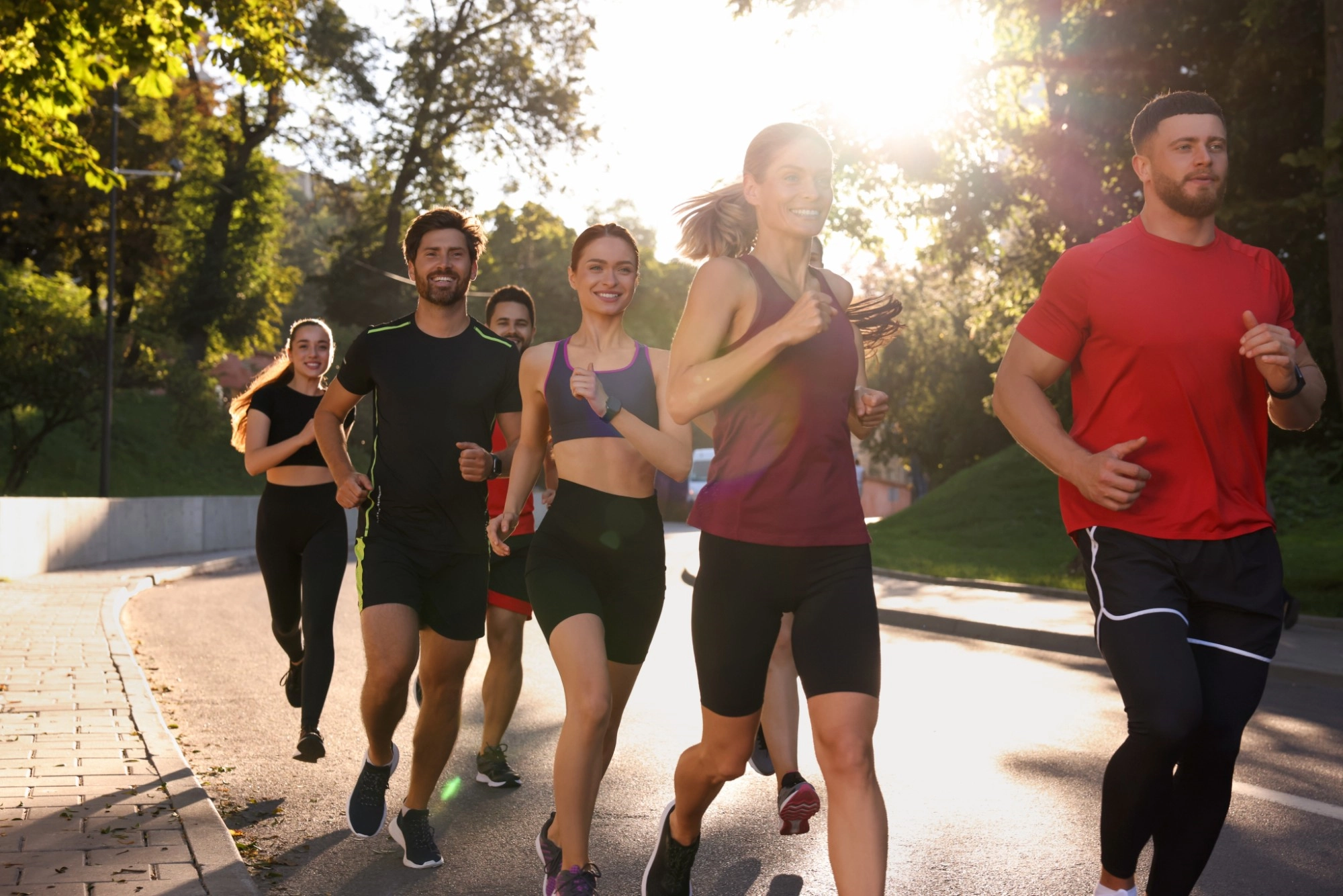 A smiling group of men and women running together along a sunlit tree-lined road