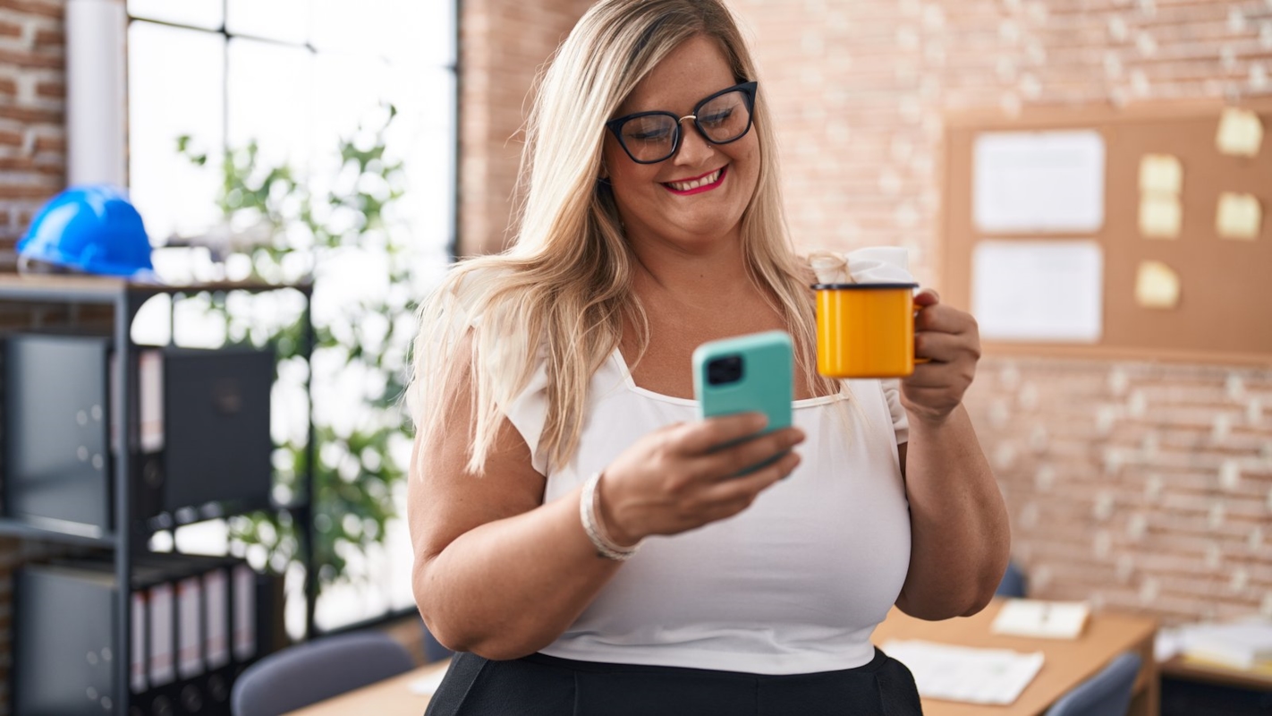 A woman smiling in an office, holding a coffee mug and her phone